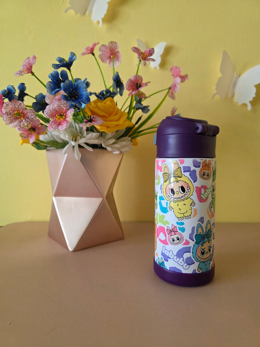Colorful water bottle with cartoon design on a yellow surface with flowers and a box.