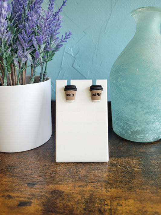 Pair of coffee-themed earrings on a white card with a blurred background of lavender and a teapot.
