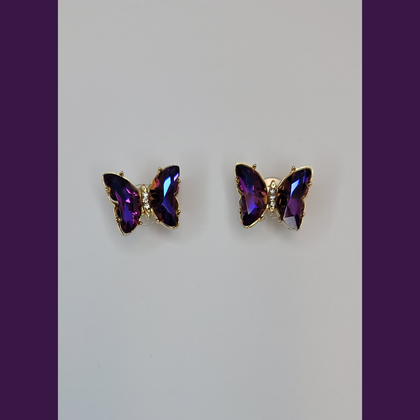 A pair of blue butterfly earrings with purple beads displayed on a white stand, with a lavender plant and a blue vase in the background.