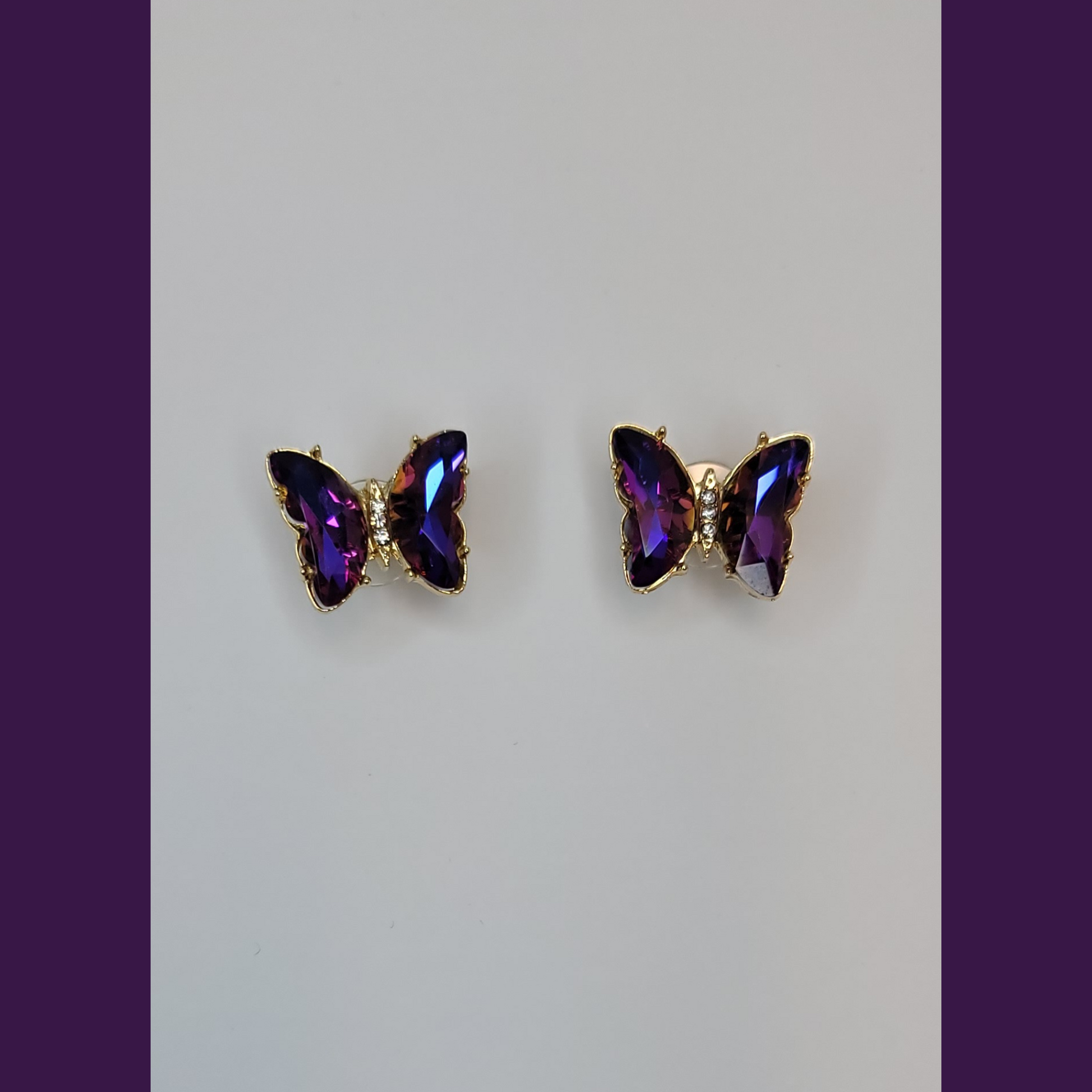 A pair of blue butterfly earrings with purple beads displayed on a white stand, with a lavender plant and a blue vase in the background.