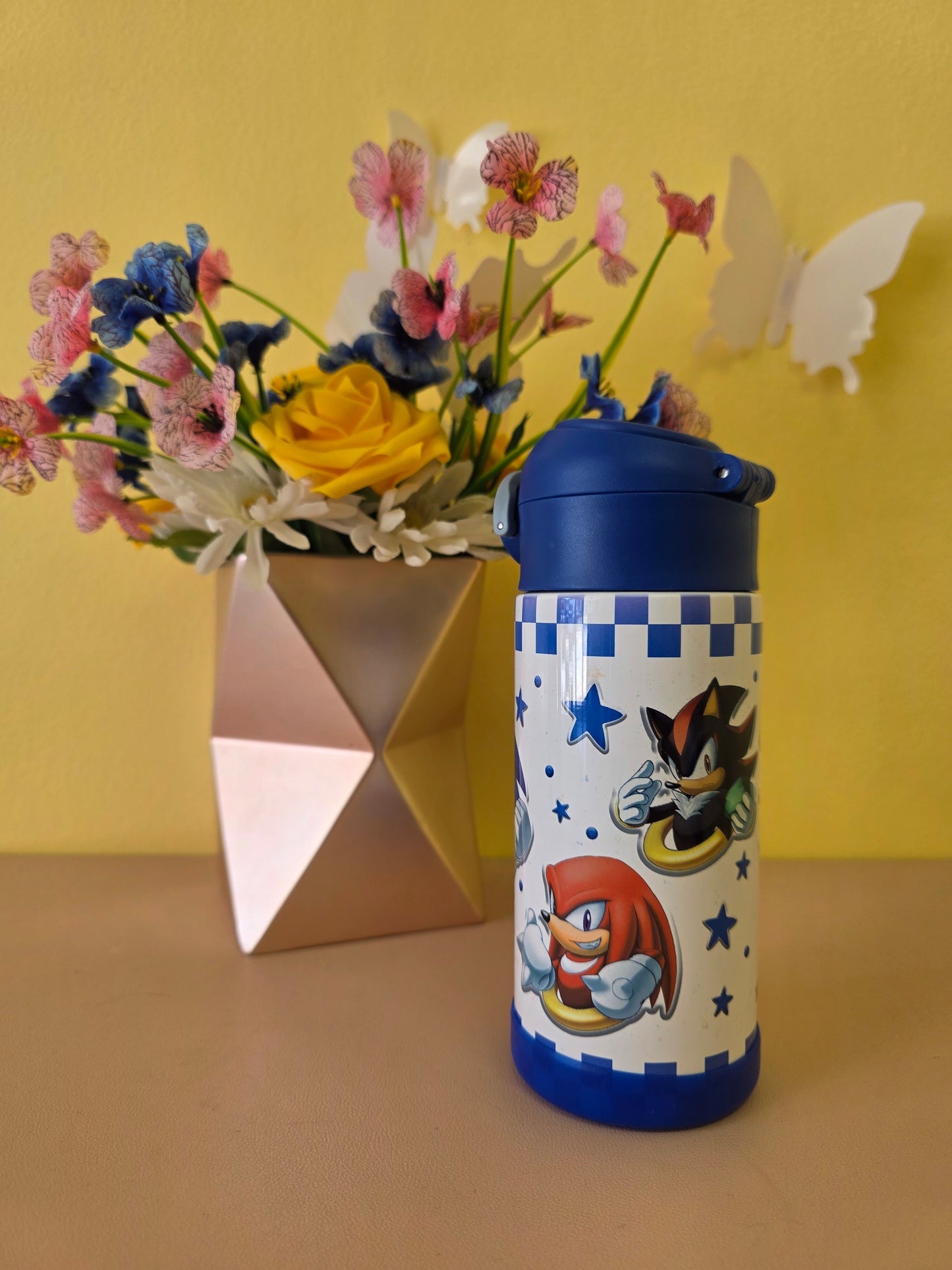 Colorful water bottle with cartoon design next to a vase of flowers on a yellow background