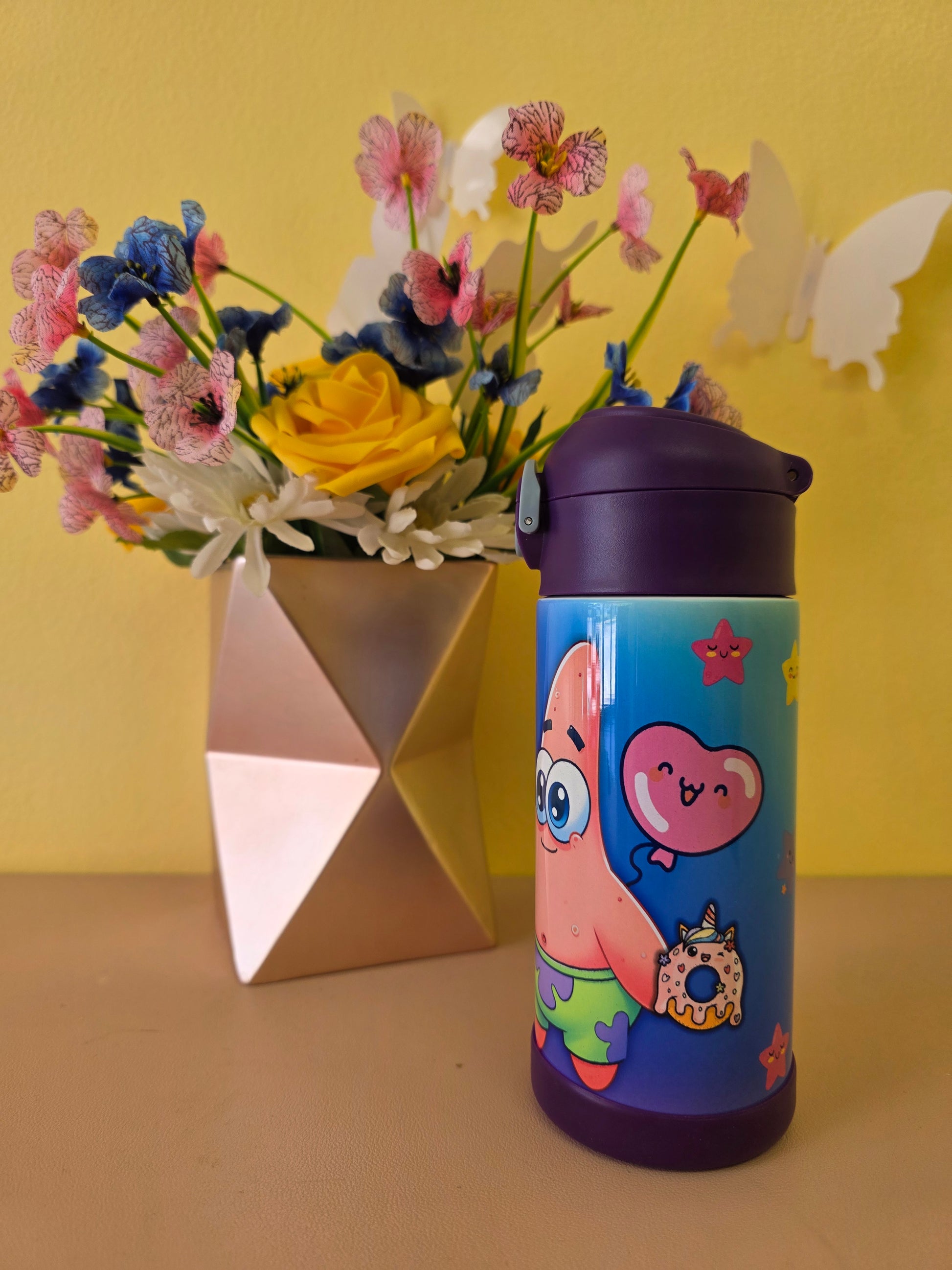 Colorful water bottle with cartoon characters next to a bouquet of flowers on a yellow surface.