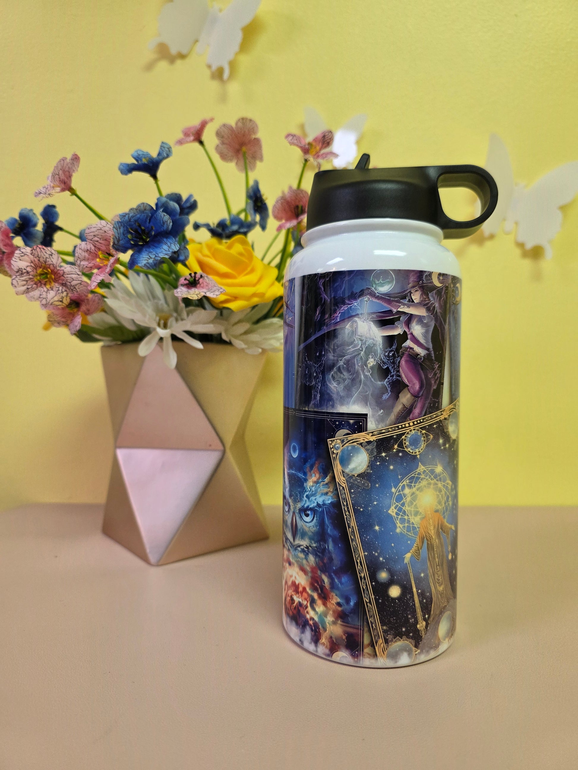 Cosmic-themed water bottle with a colorful design on a yellow surface with flowers and a box.