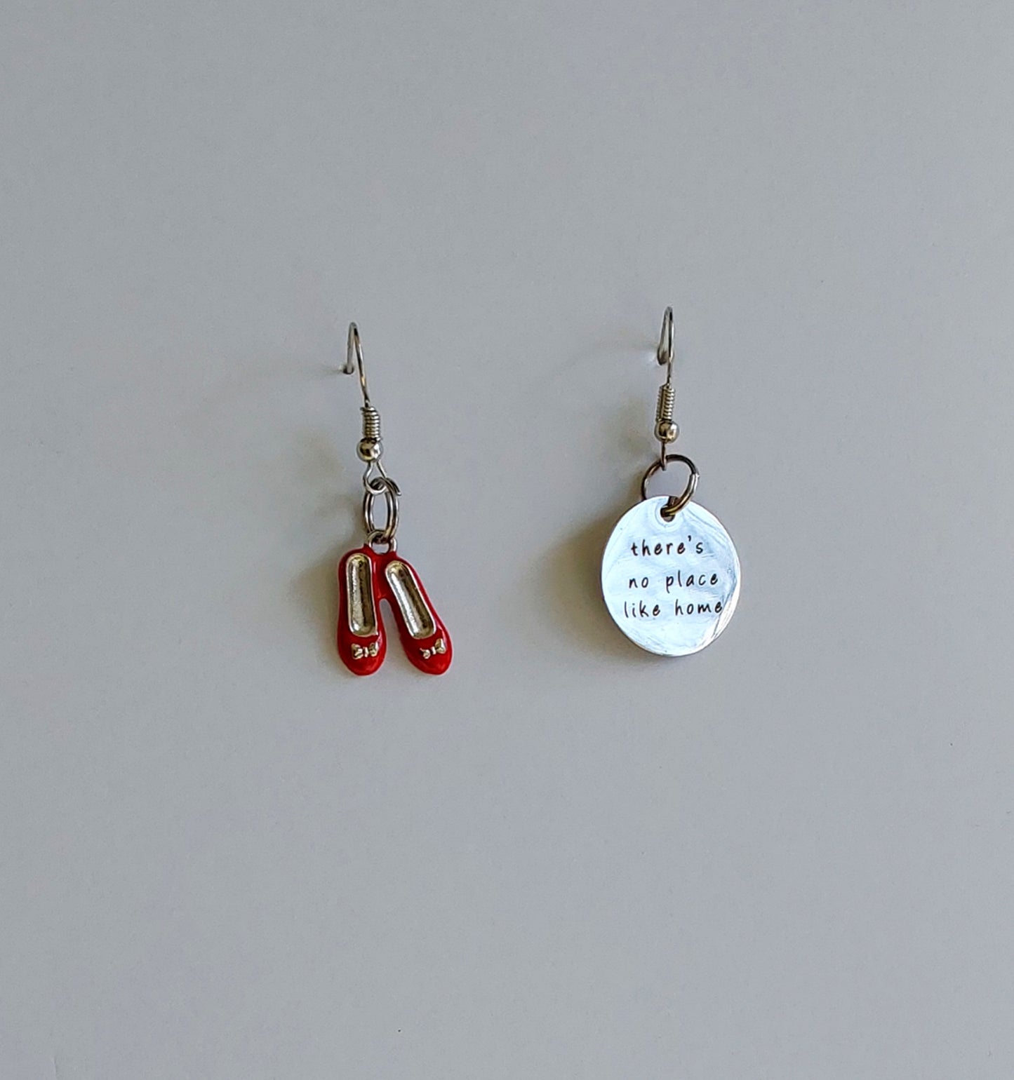 A pair of dangle earrings with one earring shaped like red slipper shoes and the other a circular charm with text on it, both on a light background.