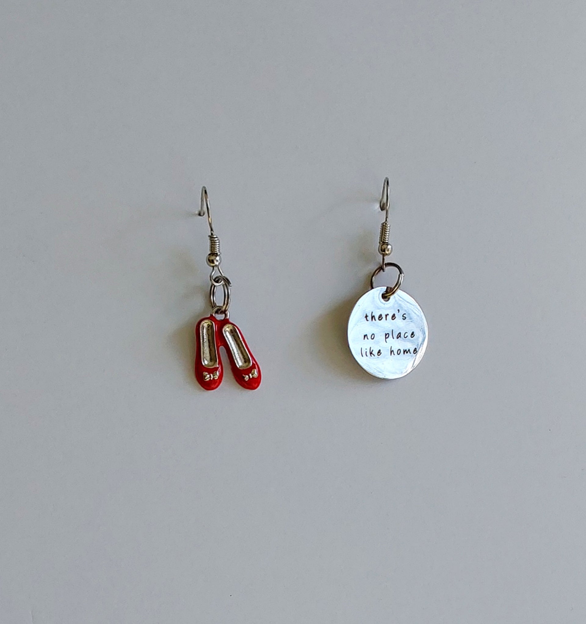 A pair of dangle earrings with one earring shaped like red slipper shoes and the other a circular charm with text on it, both on a light background.