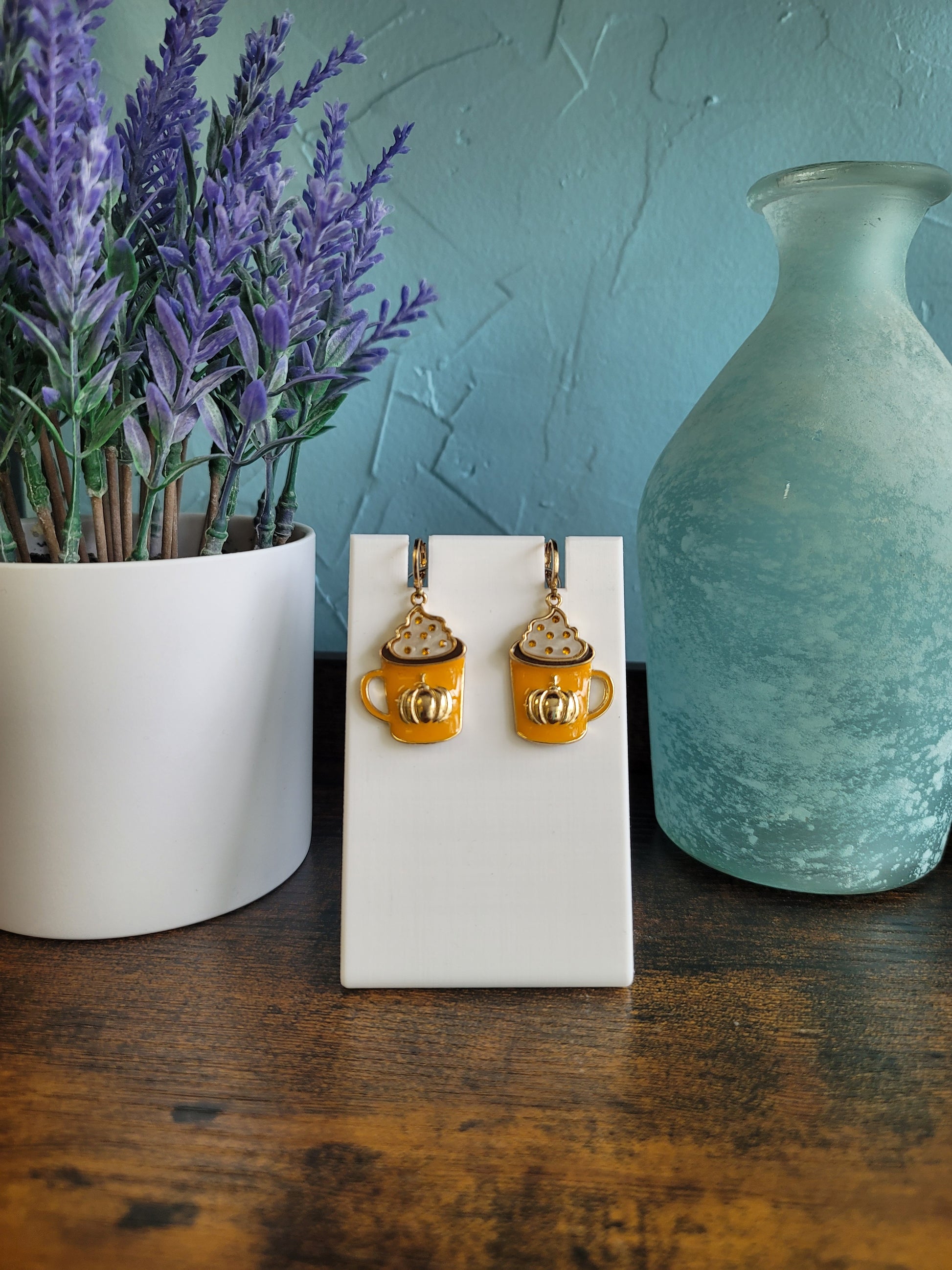 A pair of gold-colored earrings designed to resemble coffee beans, displayed on a white stand, with a lavender plant and a blue vase in the background.